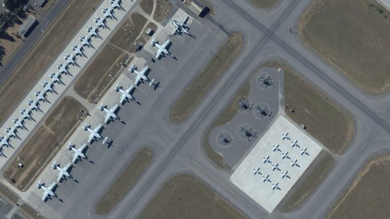 A military airfield from directly overhead, with rows of parked aircraft, helicopters and drones on a pale concrete apron.